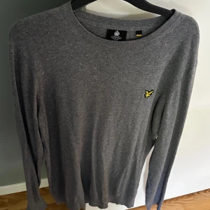 Grå tröja från Lyle & Scott - En grå långärmad tröja från Lyle & Scott med rund halsringning och ett litet gult logomärke på bröstet. Tröjan är enkel och stilren, perfekt för vardagsbruk.