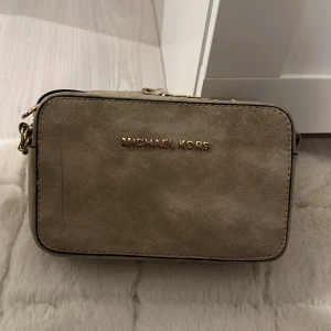 Beige axelväska från Michael Kors - En elegant beige axelväska från Michael Kors med guldfärgade detaljer och en justerbar axelrem. Väskan har en dragkedja för säker förvaring. En liten streck på väskan men inget man tänker på.