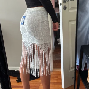 Vit spetskjol med fransar - En elegant vit spetskjol med detaljerade mönster och fransar som hänger ner från nederkanten. Kjolen är knälång och har en tight passform (AI).