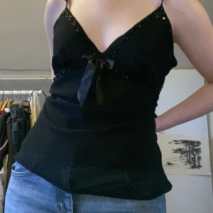 Svart sött linne - En söt svart topp med små svarta pärlor vid urringningen och en rosett i mitten. Väldigt fint skick.   OBS den är större i storlek än vad som syns på bilden. 