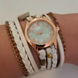 Vit och guld armbandsklocka - En elegant armbandsklocka med en vit flätad rem och gulddetaljer. Klockan har en rund urtavla med guldnummer och visare. Remmen består av flera lager, inklusive en vit flätad del, en vit slät del och en guldkedja.