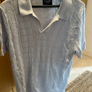 Ljusblå pikétröja från Abercrombie & Fitch - En ljusblå pikétröja från Abercrombie & Fitch med ett diskret mönster. Tröjan har korta ärmar och en klassisk krage.