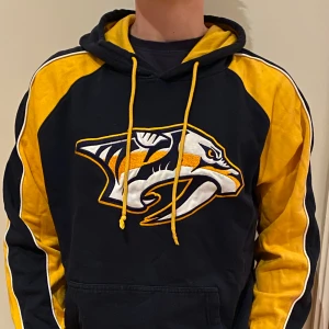 Hoodie - En vintage hoodie som är skapad av ”47”. Hoodien har ett märke med en tiger på sig. Den är mörkblå men också gul i armarna och snörena. Luvan är gul på insidan. Kvaliten är bra.