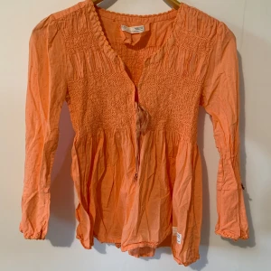 Odd Molly orange/coral blus, storlek S - En söt och romantisk blus med spetsdetaljer och en söt rosett på ärmen. Blusen har en fin smockad detalj i midjan och en v-ringning.   Skick: Mycket bra  Axelbredd: 29cm Längd: 57cm