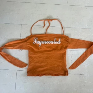 Orange topp från Jennyfer - En orange topp från Jennyfer med texten 'Sugarcoated' tryckt på framsidan. Toppen har halterneck och knytband runt ryggen! .