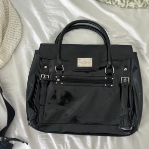 Svart handväska i lack - En elegant svart handväska i lack med två handtag och en dekorativ metallplatta på framsidan. Väskan har en dragkedja och får plats med tex en laptop.