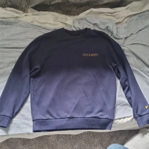 Mörkblå tröja från Lyle & Scott - En mörkblå tröja från Lyle & Scott med långärmad design. Tröjan har en rund halsringning och är dekorerad med Lyle & Scott-logotypen i gult på bröstet och vid ärmslutet.