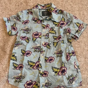 Blommig kortärmad skjorta från H&M - En färgglad kortärmad skjorta från H&M med ett blommigt mönster i lila, grönt och gult på en ljusblå bakgrund. Skjortan har knappar framtill och en klassisk krage.