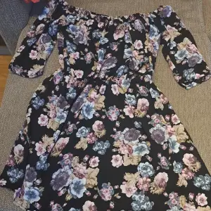 En vacker off-shoulder klänning med blommönster i olika färger som rosa, blå och beige på en svart bakgrund. Klänningen har trekvartsärmar och en elastisk midja för en bekväm passform. storlek är XS men passars S också för att det är elastisk