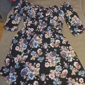 Blommig off-shoulder klänning - En vacker off-shoulder klänning med blommönster i olika färger som rosa, blå och beige på en svart bakgrund. Klänningen har trekvartsärmar och en elastisk midja för en bekväm passform. storlek är XS men passars S också för att det är elastisk