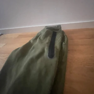 Gröna byxor med dragkedja - Ett par gröna nike tech byxor  med dragkedja på sidan. Byxorna verkar vara av ett slitstarkt material och har en avslappnad passform. köpt för 799kr är nästan helt nytt, säljer för 499kr storlek 157-170