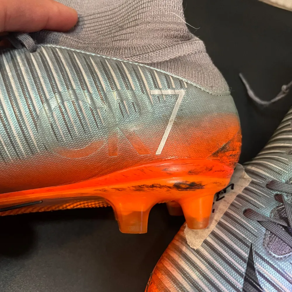 Ett par Nike Mercurial fotbollsskor i grått och orange med svarta detaljer. Skorna har snörning och en hög strumpa för extra stöd. De är märkta med 'CR7' på sidan.. Skor.