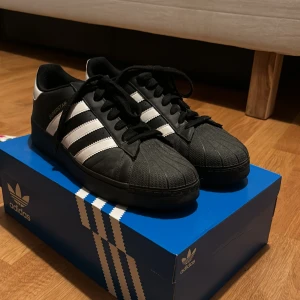 Adidas Superstar XLG originals - Ett par svarta Adidas Superstar sneakers med vita detaljer och de klassiska tre ränderna på sidorna. Skorna har snörning och Adidas-logotypen på hälen. Utöver det är skorna sparsamt använda och inga fel på skorna. Köptes från Zalando för cirka 1300kr