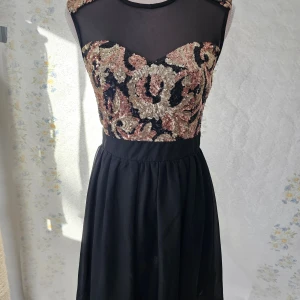 Svart klänning med paljetter - En elegant svart klänning med paljetter på överdelen. Klänningen har en transparent mesh upptill och en dragkedja på baksidan. Paljetterna är i guld och rosa nyanser och bildar ett vackert mönster. Klänningen är ärmlös och har en normal passform.