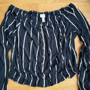 Svart offshoulder topp från H&M - Fin offshoulder topp i mjukt, luftigt material med vita ränder och knappar framtill. Toppen har långa ärmar och är elastisk i midjan ✨