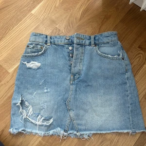 Blå jeanskjol - En blå jeanskjol med slitna detaljer och fransig nederkant. Kjolen har knappar framtill och fem fickor.