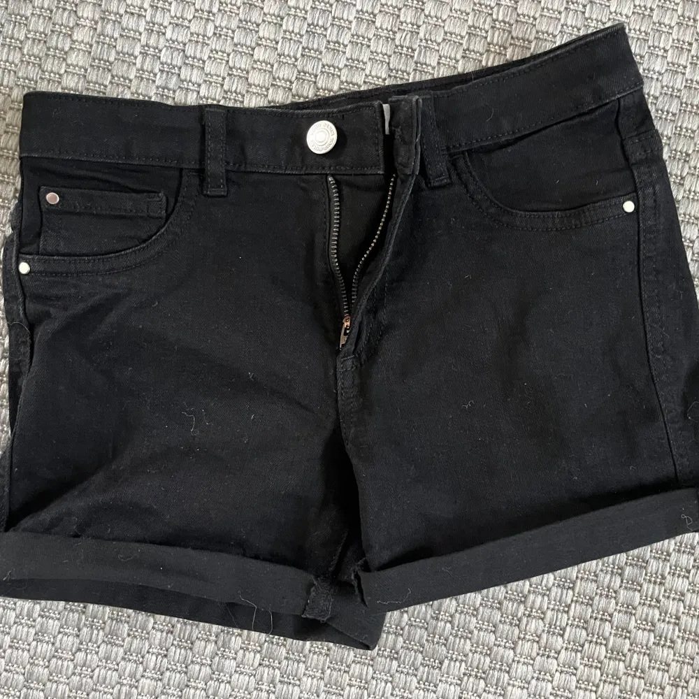 Ett par svarta jeansshorts med upprullade ben och dragkedja framtill. Shortsen har fem fickor och bälteshällor.. Shorts.