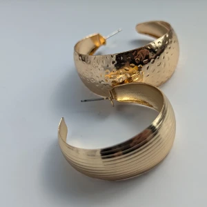 Guldfärgade örhängen med textur - Ett par guldfärgade örhängen med olika texturer. Det ena örhänget har en hamrad yta medan det andra har en räfflad yta. Båda örhängena är halvcirkelformade och har en glansig finish.