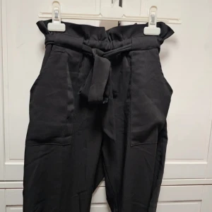 Svarta byxor från Dennis M - Svarta byxor från Dennis M i storlek S. Byxorna har en hög midja med ett knytbälte och veckade detaljer vid midjan. De har en lös passform och är tillverkade i ett mjukt material.