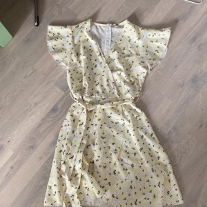 Ljusgul blommig klänning med volanger - Säljer en superfin ljusgul klänning med små blommor i svart och vitt. Jag upplever att klänningen är lite mer gul i verkligheten än vad den är på bilderna. Klänningen har korta ärmar med volanger och ett knytband i midjan för en fin passform. Den är gjord i ett mjukt och lätt material, perfekt för sommar och vår. Klänningen har en dragkedja i ryggen för enkel på- och avklädning. Perfekt för fest eller en dag ute i solen!