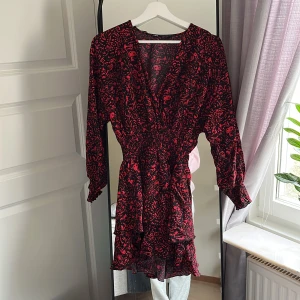 Svart och röd klänning från Zara - Säljer en superfin svart och röd klänning från Zara i storlek XS. Klänningen har ett blommigt mönster och är perfekt för festliga tillfällen. Den har långa ärmar och en v-ringad hals. Klänningen är i nyskick och har en volangdetalj längst ner. Passformen är normal och materialet känns mjukt och skönt.