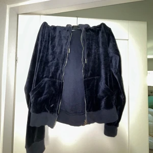 Svart velourjacka från Juicy Couture - Säljer en snygg och bekväm svart velourjacka från Juicy Couture. Jackan har en dragkedja framtill och två fickor. Den är långärmad och har en huva. Perfekt för en avslappnad och stilren look!
