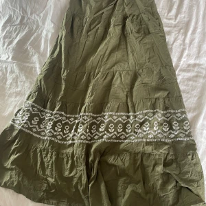 Grön långklänning med broderad detalj - Säljer en superfin grön långkjol med en vacker vit broderad detalj runt nederdelen. Den är perfekt för sommaren och är superbekväm att bära. Materialet känns lätt och luftigt, bra för varma dagar. Den är i nyskick och redo att bli en del av din garderob! Kjolen kan även användas som en klänning då man knyter bandet bakom nacken båda har funkat lika bra💖