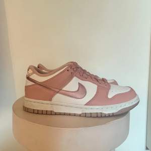 Säljer ett par snygga Nike Dunk sneakers i rosa och vitt. Skorna har en klassisk design med rosa läderdetaljer och vit bas. De har snörning framtill och Nike-loggan på hälen i rött. Perfekta för vardagsbruk eller att ge din outfit en sportig touch!