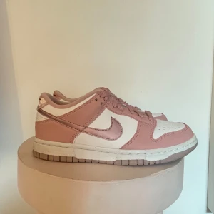 Nike Dunk sneakers i rosa och vitt - Säljer ett par snygga Nike Dunk sneakers i rosa och vitt. Skorna har en klassisk design med rosa läderdetaljer och vit bas. De har snörning framtill och Nike-loggan på hälen i rött. Perfekta för vardagsbruk eller att ge din outfit en sportig touch!