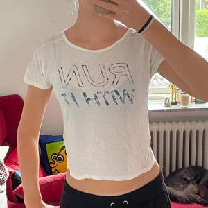 Säljer en vit croppad t-shirt med texten 'RUN WITH IT' i ett coolt mönster. T-shirten är kortärmad och perfekt för sommarens varma dagar. Den är i bra skick och passar perfekt för träning eller en avslappnad dag ute. 🏃‍♀️