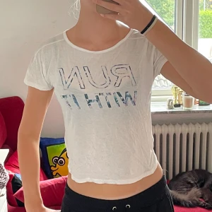 Vit croppad t-shirt med text - Säljer en vit croppad t-shirt med texten 'RUN WITH IT' i ett coolt mönster. T-shirten är kortärmad och perfekt för sommarens varma dagar. Den är i bra skick och passar perfekt för träning eller en avslappnad dag ute. 🏃‍♀️