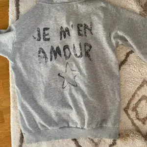 En grå hoodie från Gina Tricot med texten 'JE M'EN AMOUR' och en stjärna tryckt på både framsidan och baksidan. Hoodien har långa ärmar och ribbstickade muddar vid ärmslut och nederkant. har tyvärr blivit lite nopprig!☺️