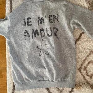 Grå hoodie från Gina Tricot - En grå hoodie från Gina Tricot med texten 'JE M'EN AMOUR' och en stjärna tryckt på både framsidan och baksidan. Hoodien har långa ärmar och ribbstickade muddar vid ärmslut och nederkant. har tyvärr blivit lite nopprig!☺️