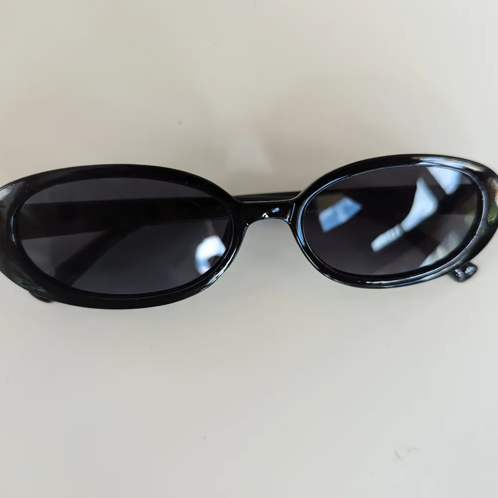 Ett par svarta ovala solglasögon med mörka linser. Glasögonen har en slät och blank yta med en enkel och stilren design.. Accessoarer.