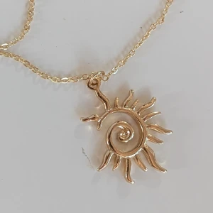 Guldhalsband med solhänge - Ett elegant guldhalsband med ett solformat hänge. Hänget har en spiralformad mitt och strålar som sträcker sig utåt, vilket ger det ett unikt och konstnärligt utseende. Kedjan är tunn och matchar hängets guldiga färg.