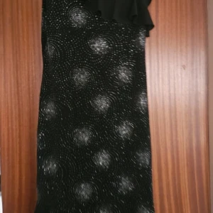 Svart glittrig klänning med volangdetalj - Snygg svart klänning med glittrigt mönster och volangdetalj vid axeln. Klänningen har en elegant passform och är perfekt för festliga tillfällen. Den har en smickrande längd som slutar vid knäna. Står inte vad det är för stl. Jag har M 38 och den passar mig