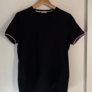 Svart t-shirt från Moncler - Snygg svart t-shirt från Moncler med slim fit passform. Den har korta ärmar med röd och vit randdetalj samt Moncler-logga på ärmen. Perfekt för en stilren look.