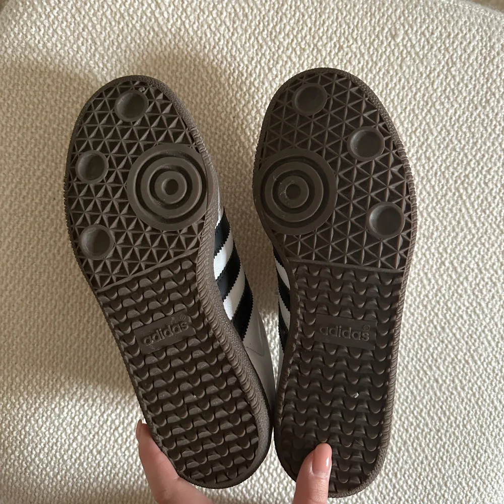 Klassiska Adidas Samba sneakers med vit ovandel, grå mockadetaljer och svarta ränder på sidorna. Perfekta för en sportig och stilren look. Storlek 36 2/3 - passar mig med 37. Använda 2-3 gånger och inköpta förra året. 🖤. Kengät.