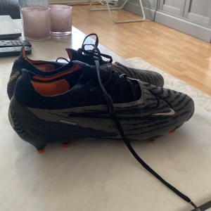 Nike Phantom GX fotbollsskor i svart och grå Elite versionen - Säljer ett par Nike Phantom GX fotbollsskor i svart och grå med orange detaljer. Skorna har snörning och mönstrad ovandel för extra grepp på planen. Perfekta för dig som vill ha stilrena och funktionella fotbollsskor.
