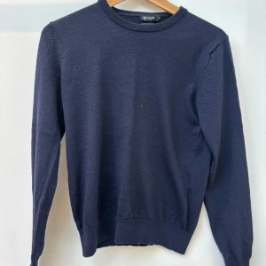 Mörkblå ulltröja från Tiger of Sweden - Säljer en klassisk mörkblå tröja från Tiger of Sweden i 100% ull. Tröjan har rund halsringning, långa ärmar och ribbade muddar vid ärmslut och nederkant. Perfekt för kyligare dagar och enkel att matcha med olika outfits.