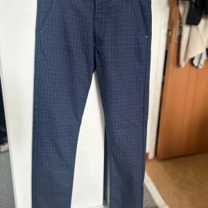 Mörkblå rutiga byxor från Hawli's JNS - Snygga mörkblå byxor från Hawli's JNS med  rutigt mönster. Byxorna har normal passform, raka ben och är tillverkade i 100% bomull! Perfekta för dig som letar efter ett par stiliga kostymbyxor till ditt nästa event!