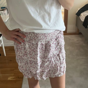 Vit blommig volangkjol från BikBok - Supersöt kort kjol från BikBok i vitt med små rosa blommor och flera volanger. Kjolen har resår i midjan och är perfekt för en somrig look. Lätt och luftig, passar perfekt till en enkel topp.