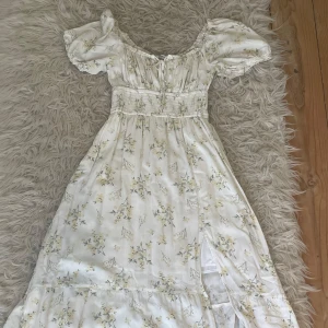 Vit blommig långklänning med puffärmar - Romantisk långklänning i vit med små blommor i gult och grönt. Klänningen har puffärmar, smock i midjan och en slits framtill. Även en söt knytning vid bysten. Perfekt för sommarens alla tillfällen.
