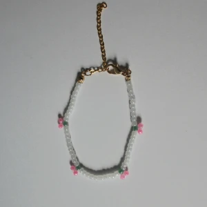 Pärlarmband med små rosa blommor, bracelet  - Sött armband med vita pärlor och små rosa blommor, samt gröna detaljer. Armbandet har guldfärgat justerbart spänne och passar perfekt som en färgglad accessoar till dig själv eller som en present. 