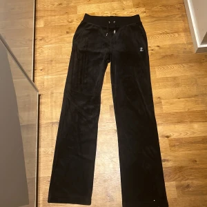 Svarta velourbyxor från Juicy Couture - Säljer ett par klassiska svarta velourbyxor från Juicy Couture med strassdetaljen 'Juicy' på baksidan. Aldrig använt! Byxorna har dragsko i midjan och raka ben. Perfekta för en avslappnad och trendig look.