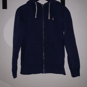 Mörkblå hoodie från Polo Ralph Lauren - Mörkblå hoodie från Polo Ralph Lauren med dragkedja och huva. Tröjan har vita snören vid huvan, två fickor framtill och en liten gul broderad logga på bröstet. Perfekt för en avslappnad stil.