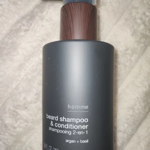 Beard Shampoo & Conditioner 2-i-1 - Säljer en 2-i-1 skäggschampo och balsam från Homme med argan och basilika. Perfekt för att hålla skägget mjukt och fräscht. Flaskan har en stilren design med en träliknande pump.