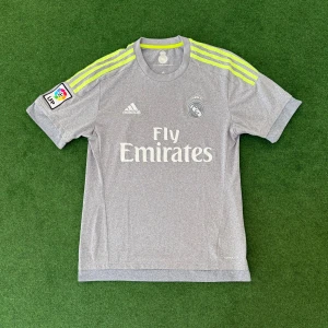 Grå Real Madrid fotbollströja från Adidas - Säljer en grå Real Madrid fotbollströja med neongula detaljer och Adidas-logga. Tröjan har korta ärmar, La Liga-märke på ena ärmen och Climacool-material för extra komfort. Klassisk Real Madrid-logga och Fly Emirates-tryck på bröstet.