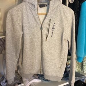 Grå hoodie från Sail Racing - Säljer en stilren grå hoodie från Sail Racing med dragkedja och en praktisk bröstficka. Hoodien har en broderad logga på bröstet och en på huvan. Den är skön,varm och snygg. Perfekt för en lite kylig sommarkväll.