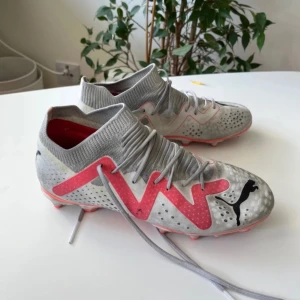Grå och rosa fotbollsskor från Puma - Snygga fotbollsskor från Puma i grått och rosa med svarta detaljer. Skorna har snörning och en rund tå, perfekt för fotbollsplanen. De har en modern design med mönstrade detaljer och en slitstark sula för bra grepp.